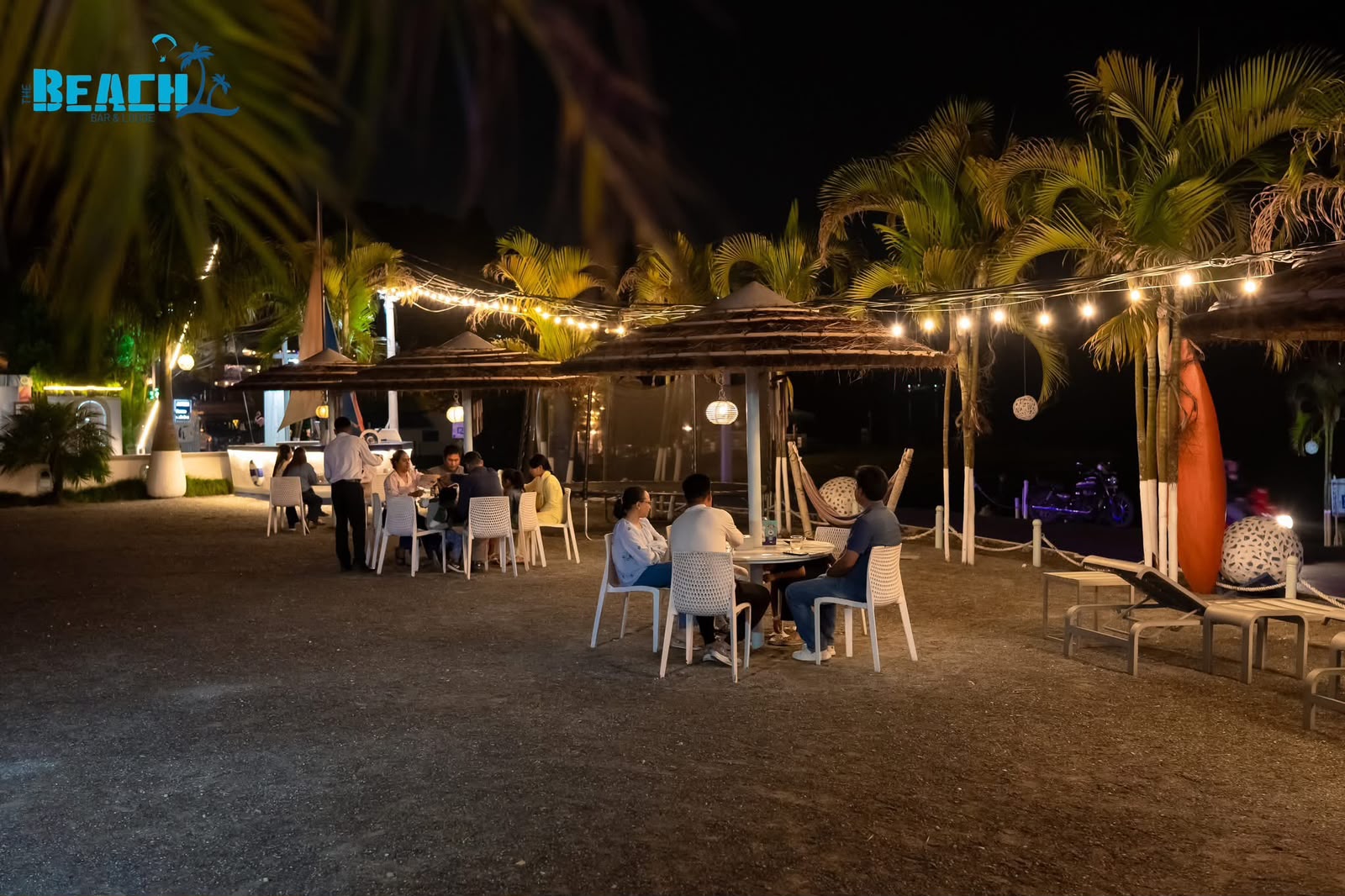 Lakeside tables beneath warm string lights among palms at night