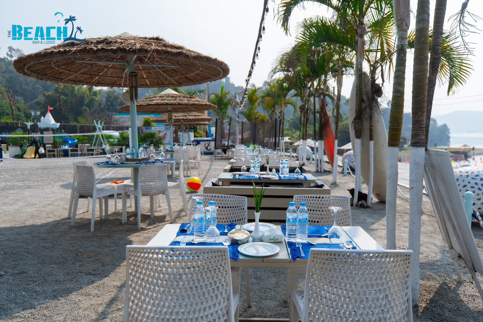 White dining tables beneath thatched parasols and palms, beside Phewa Lake